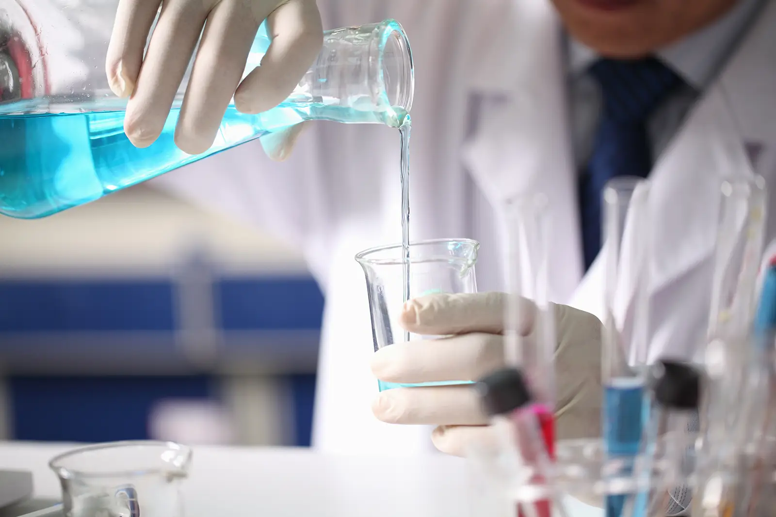 A person wearing a lab coat and gloves pouring a blue liquid from a flask into a beaker in a laboratory setting.