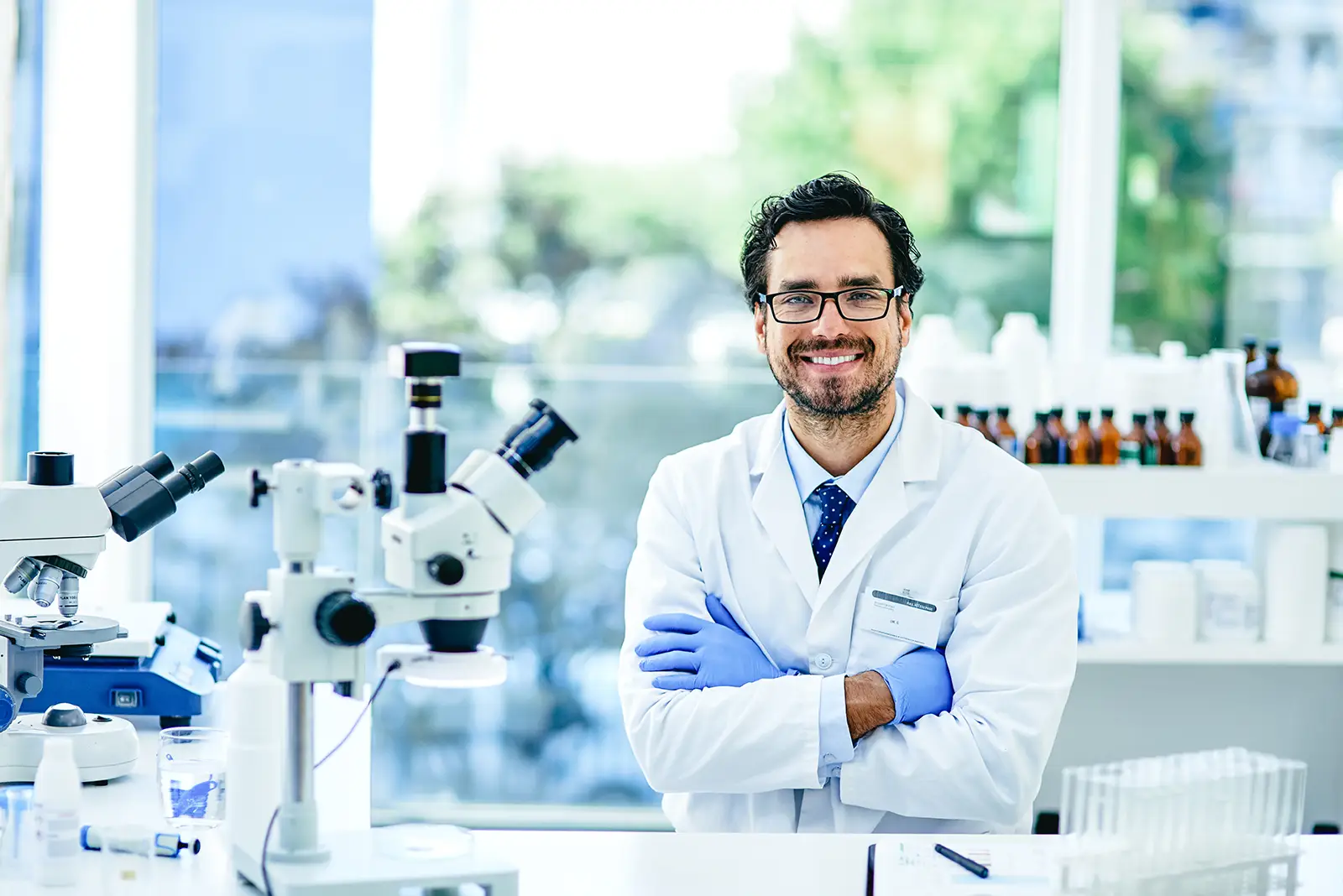 A scientist in a white lab coat and blue gloves stands in a research chemical laboratory, with microscopes and glassware on the table in front of a bright, windowed background.
