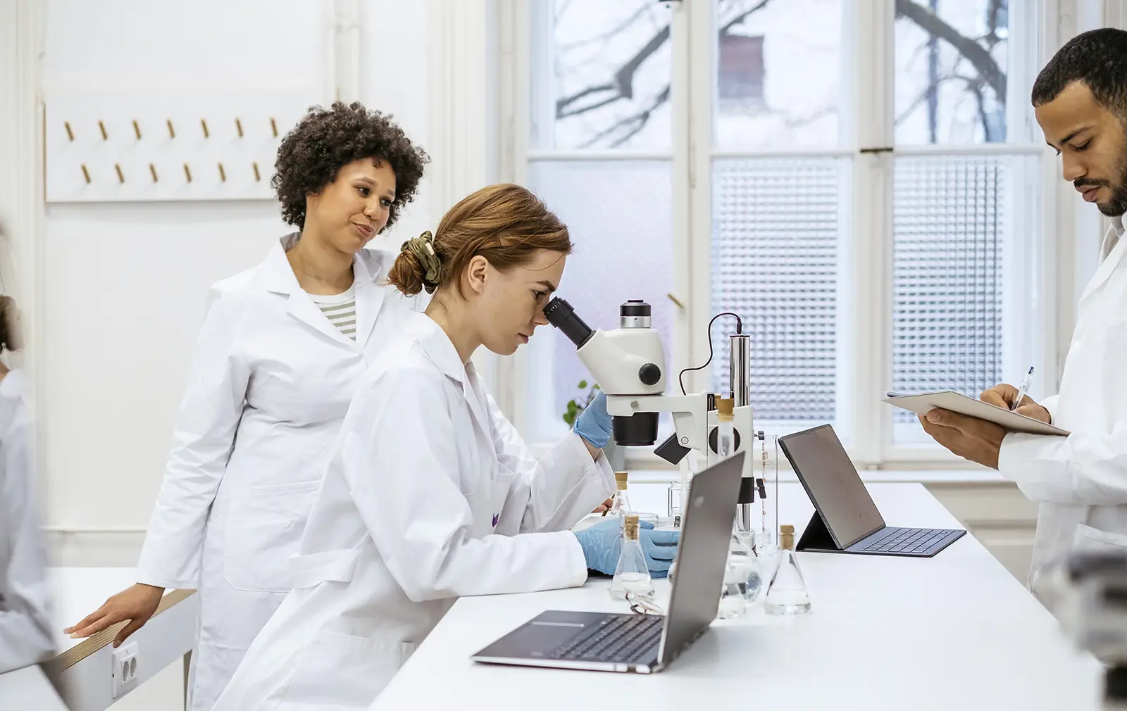 Researchers in a laboratory dressed in white coats; one is using a microscope, another is writing notes, and a third person is observing.