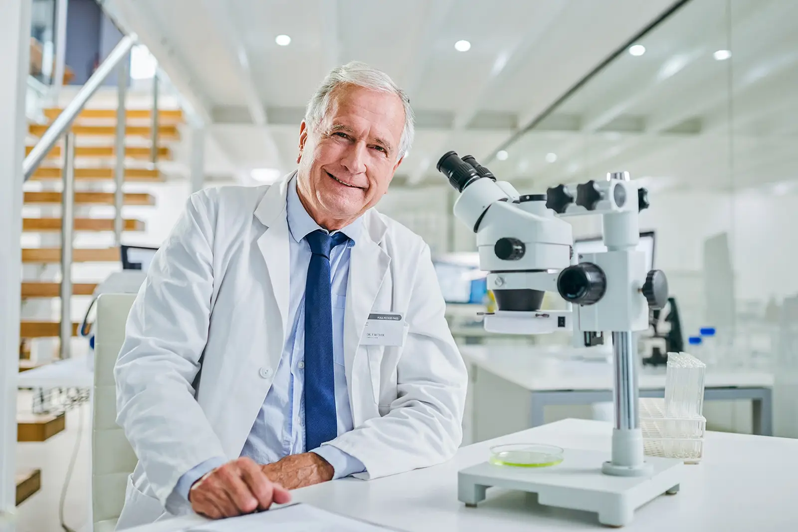 An older man in a white lab coat sitting at a desk in a research laboratory, with a microscope and test tubes nearby, smiling at the camera.