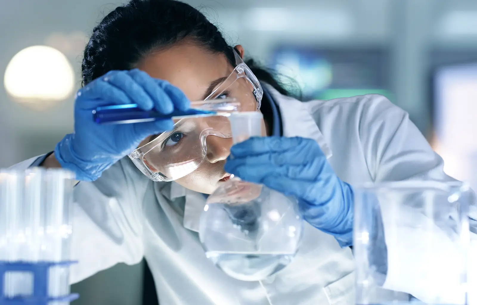 A scientist wearing safety goggles and gloves carefully pours liquid from a test tube into a flask in a laboratory setting for illustrative purposes only
