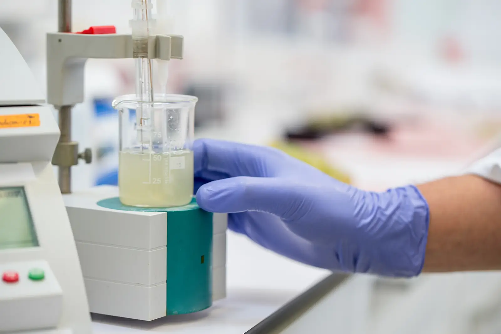 A gloved hand holding a beaker filled with a yellow liquid, placed on a lab instrument in a laboratory setting.