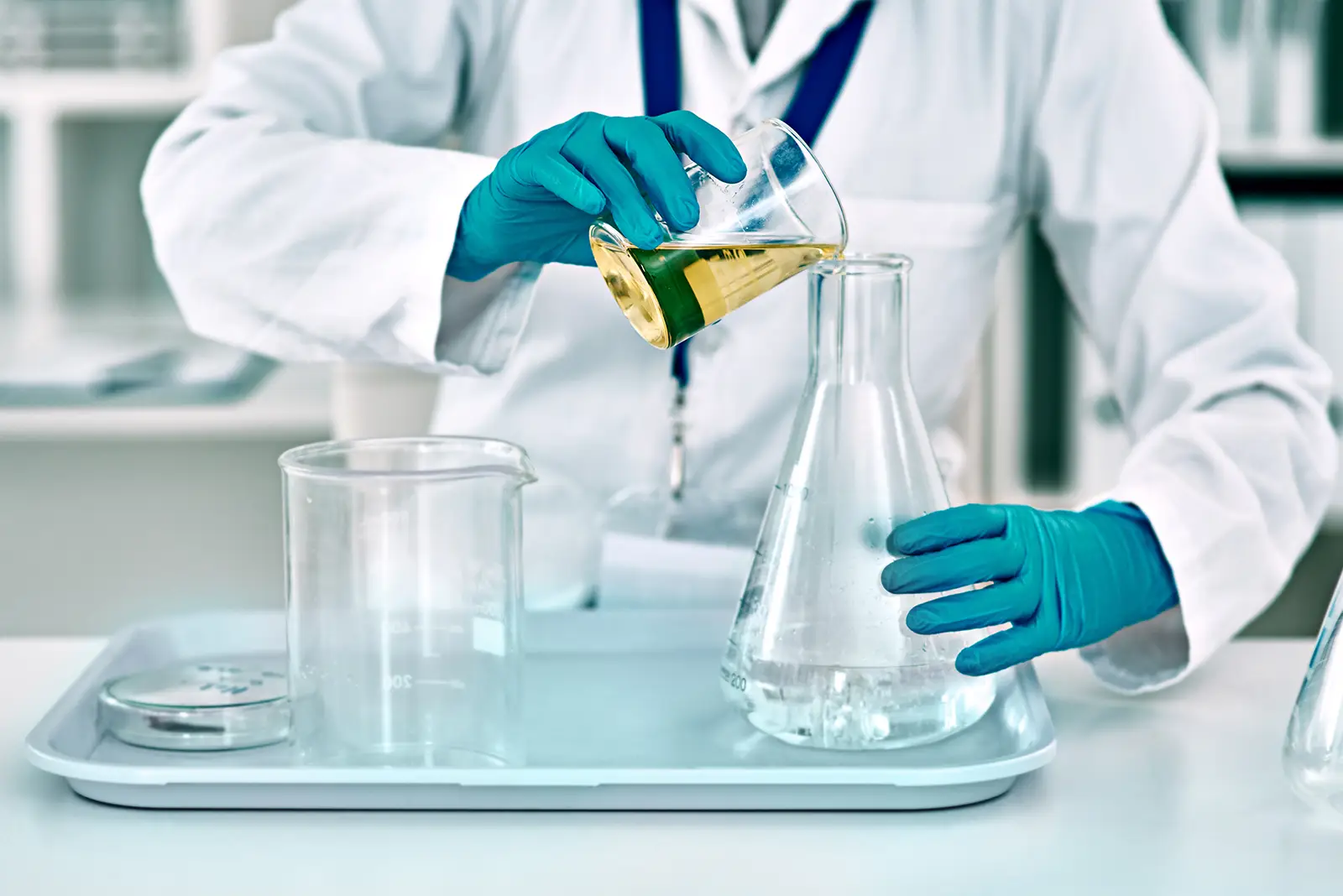 A person in a lab coat and gloves pours a yellow liquid from a beaker into a flask in a laboratory setting.