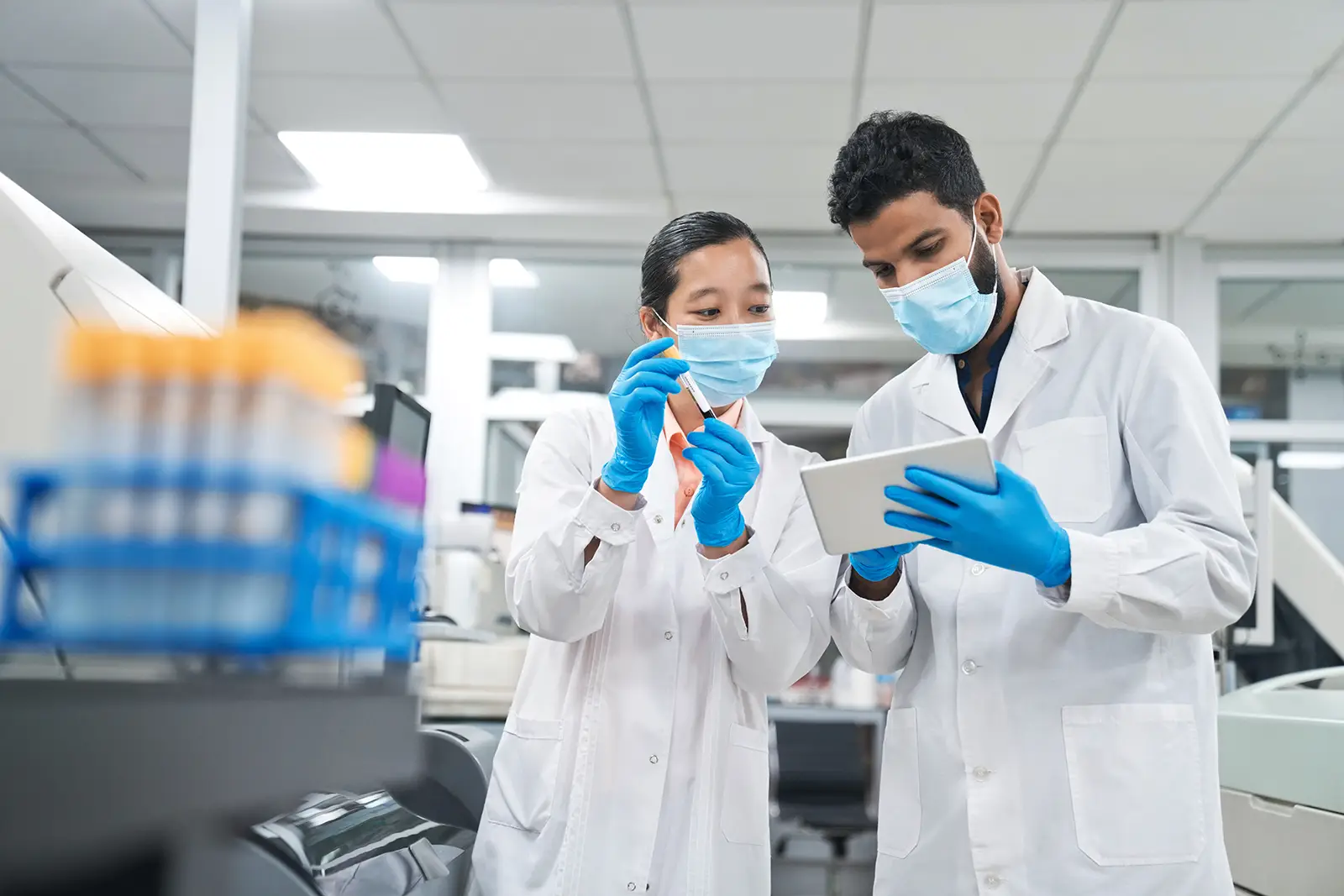 Two scientists wearing lab coats, masks, and gloves, examining a sample and discussing data on a tablet in a laboratory setting.