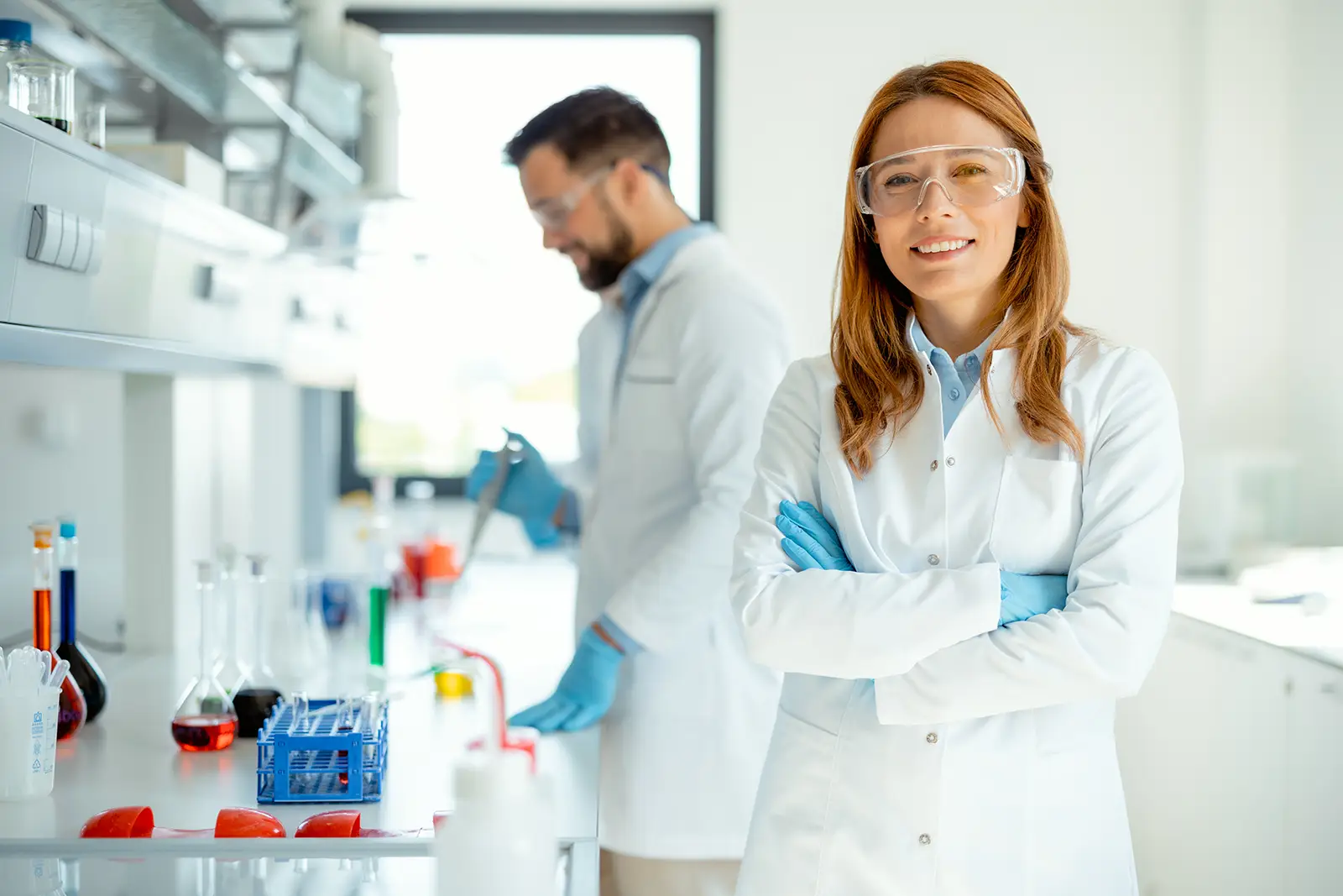 Two scientists wearing lab coats and safety goggles working in a laboratory, with one woman smiling in the foreground and a man using a pipette in the background, surrounded by various glassware and laboratory equipment.