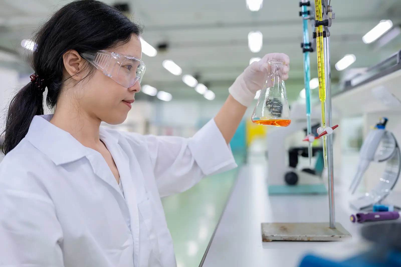 A researcher in a laboratory wearing protective eyewear and gloves, holding a flask with orange liquid, with lab equipment in the background.